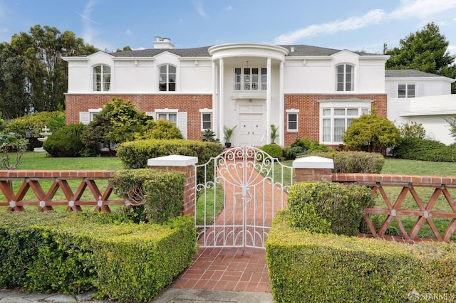 greek revival inspired property with a fenced front yard, a gate, brick siding, and stucco siding