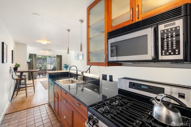 kitchen with stainless steel appliances, a sink, brown cabinetry, dark countertops, and glass insert cabinets