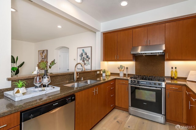 kitchen with under cabinet range hood, dark stone countertops, a sink, arched walkways, and appliances with stainless steel finishes