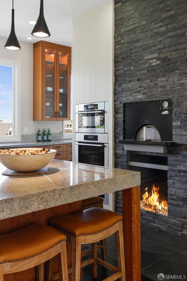 kitchen with light stone counters, a stone fireplace, a kitchen breakfast bar, brown cabinetry, and stainless steel double oven