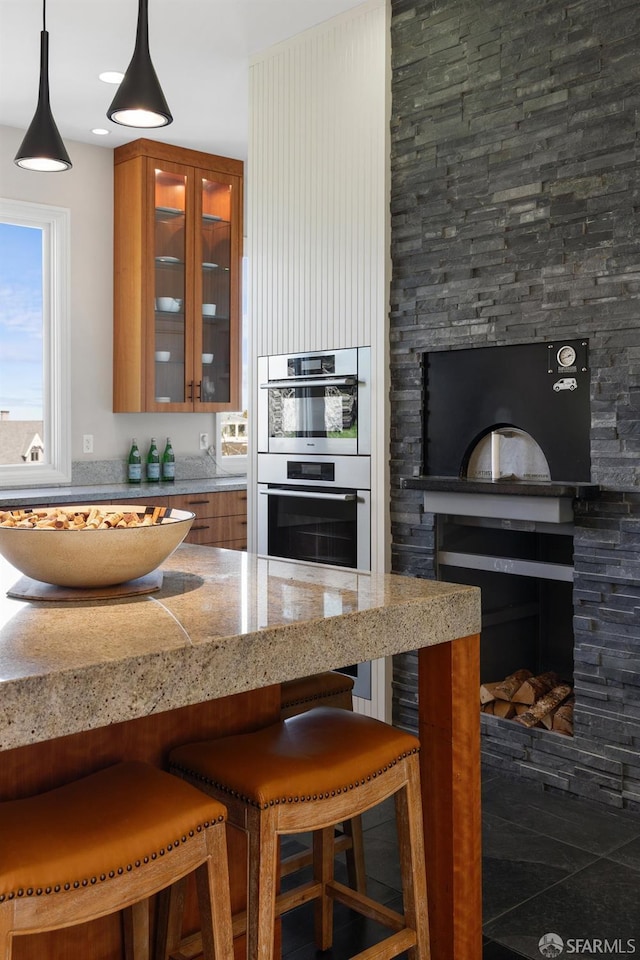 kitchen with a breakfast bar, light stone counters, double oven, brown cabinetry, and glass insert cabinets