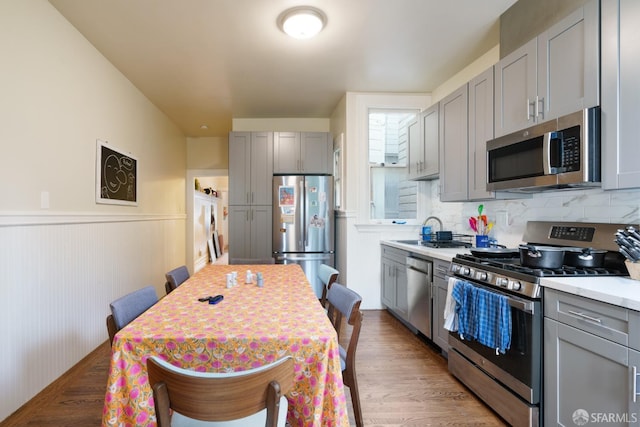 kitchen with light wood finished floors, a wainscoted wall, gray cabinets, stainless steel appliances, and light countertops