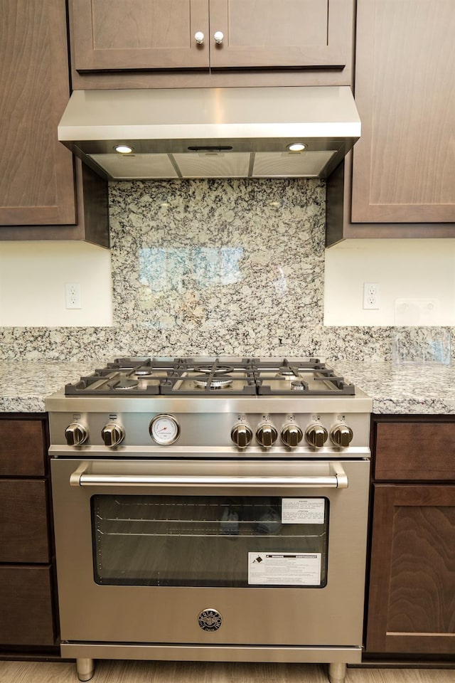 kitchen with exhaust hood, dark brown cabinets, stainless steel range, light stone counters, and tasteful backsplash