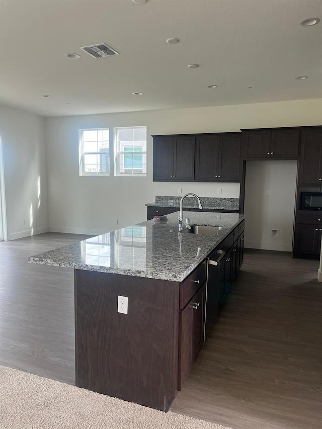 kitchen with sink, a kitchen island with sink, light stone counters, dark brown cabinets, and built in microwave