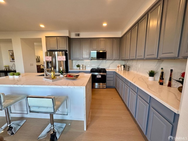 kitchen with appliances with stainless steel finishes, gray cabinetry, backsplash, a kitchen island with sink, and light hardwood / wood-style flooring