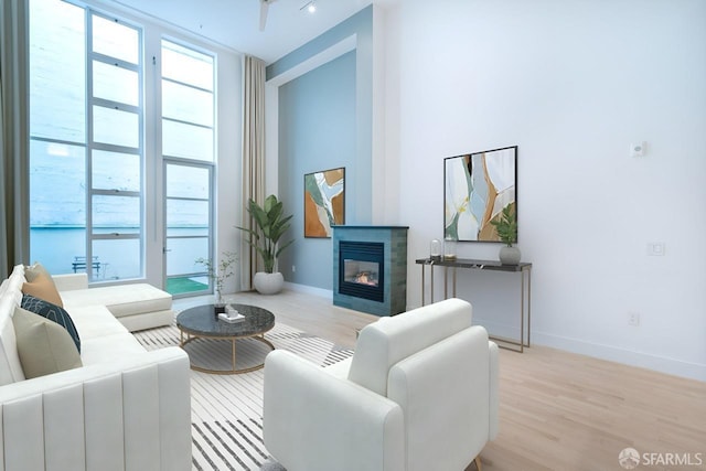 living area featuring baseboards, a glass covered fireplace, a towering ceiling, light wood-style flooring, and a wall of windows