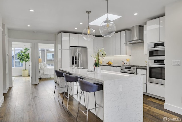 kitchen featuring white cabinets, an island with sink, stainless steel appliances, and wall chimney range hood