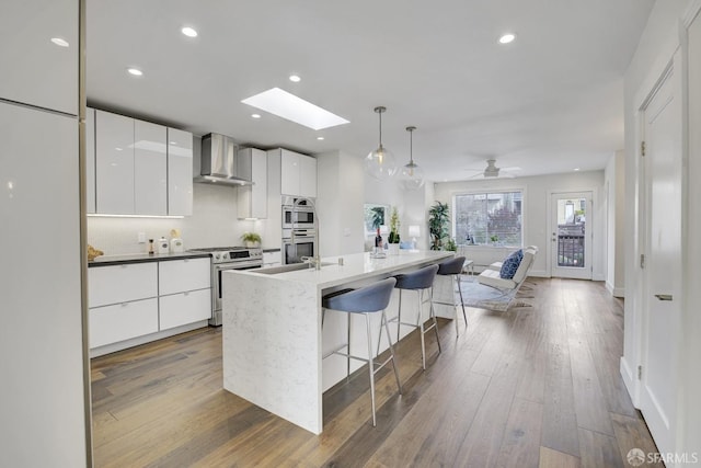 kitchen featuring a skylight, white cabinetry, stainless steel appliances, and wall chimney range hood