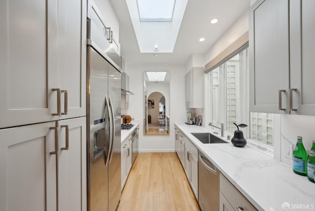 kitchen featuring appliances with stainless steel finishes, a skylight, sink, light stone countertops, and light hardwood / wood-style flooring