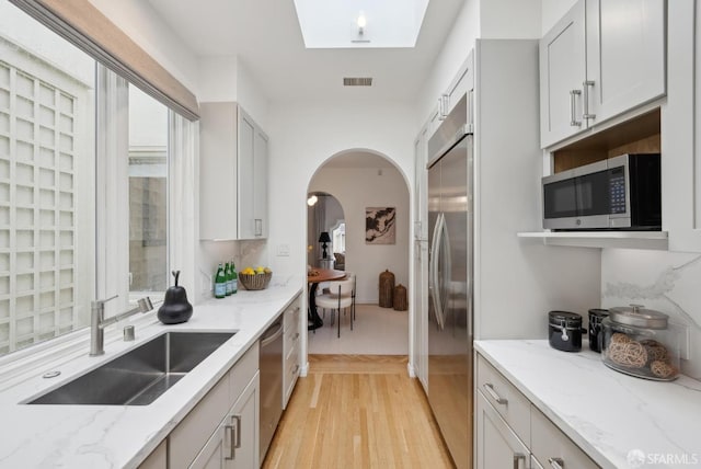 kitchen featuring sink, appliances with stainless steel finishes, a skylight, light stone countertops, and light wood-type flooring