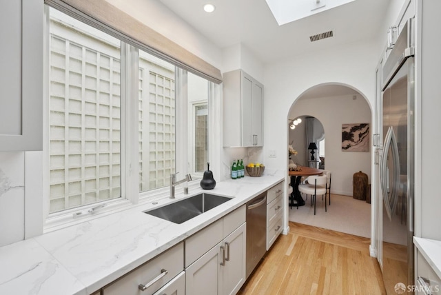 kitchen featuring a skylight, sink, stainless steel appliances, light stone countertops, and light hardwood / wood-style flooring