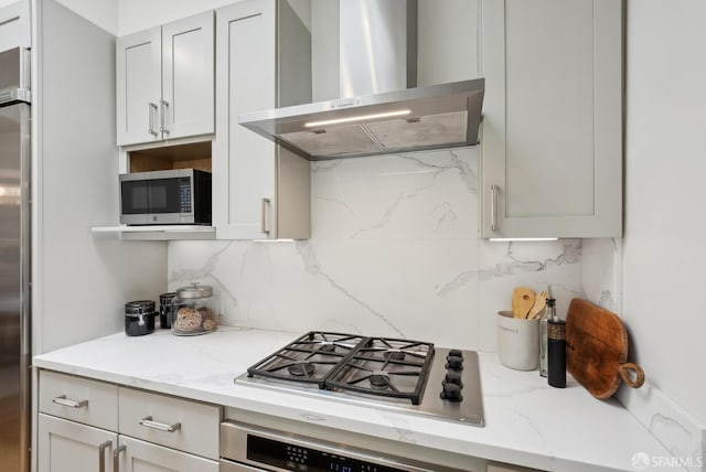 kitchen with wall chimney range hood, decorative backsplash, light stone countertops, and appliances with stainless steel finishes