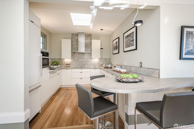 kitchen featuring hanging light fixtures, wall chimney range hood, a kitchen bar, a skylight, and white cabinetry