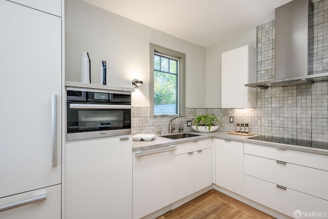 kitchen featuring wall chimney range hood, oven, black electric stovetop, white cabinetry, and sink