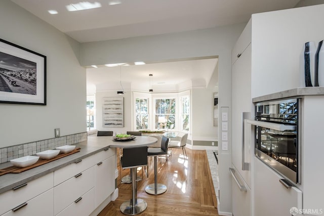 kitchen with white cabinets, stainless steel oven, light wood-type flooring, kitchen peninsula, and decorative backsplash