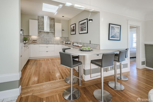 kitchen featuring kitchen peninsula, wall chimney exhaust hood, decorative light fixtures, white cabinetry, and a kitchen breakfast bar