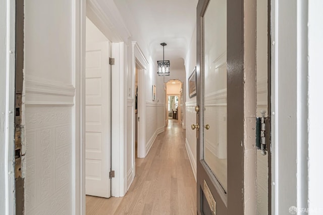 hallway with arched walkways, a decorative wall, wainscoting, and light wood finished floors