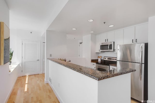 kitchen with stainless steel appliances, dark stone countertops, kitchen peninsula, white cabinets, and light wood-type flooring