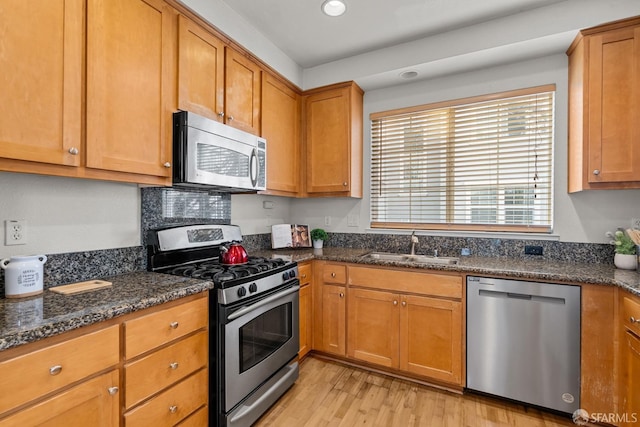 kitchen featuring dark stone countertops, light wood-type flooring, a sink, stainless steel appliances, and brown cabinets