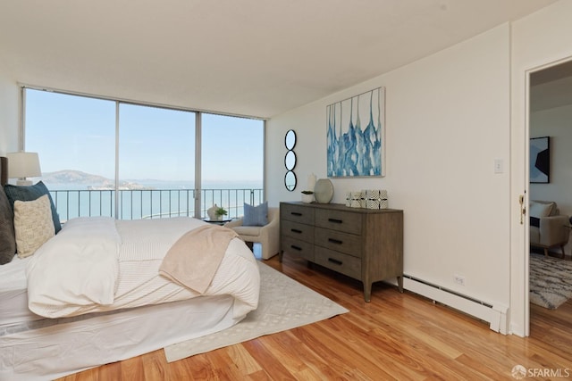 bedroom featuring access to exterior, a baseboard heating unit, a water view, light hardwood / wood-style flooring, and a wall of windows