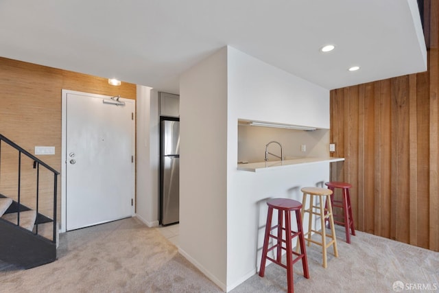 kitchen with light colored carpet, freestanding refrigerator, wood walls, a sink, and a kitchen breakfast bar