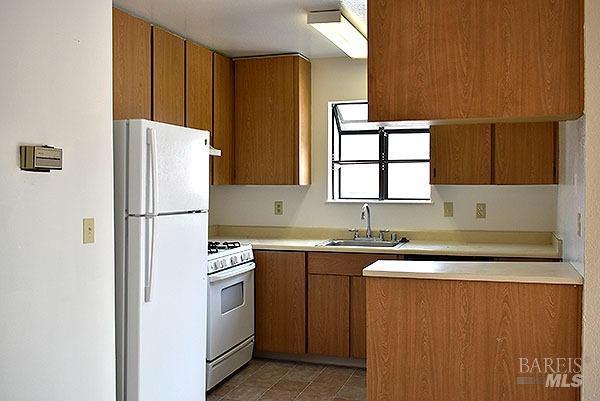 kitchen featuring tile patterned floors, a sink, white appliances, brown cabinetry, and light countertops