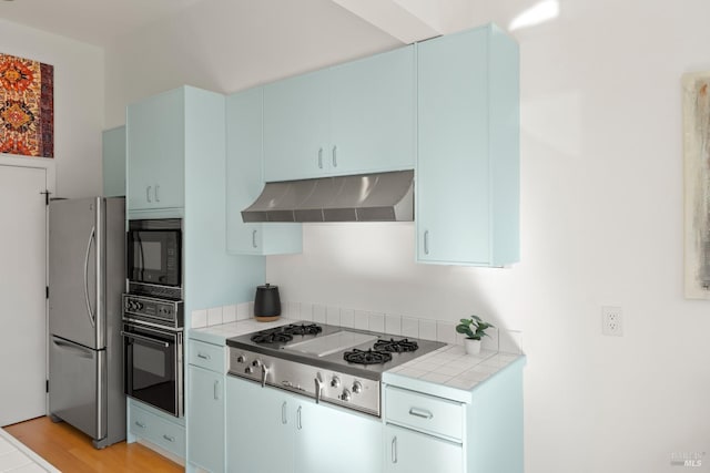 kitchen featuring tile counters, light hardwood / wood-style floors, black appliances, blue cabinetry, and wall chimney exhaust hood