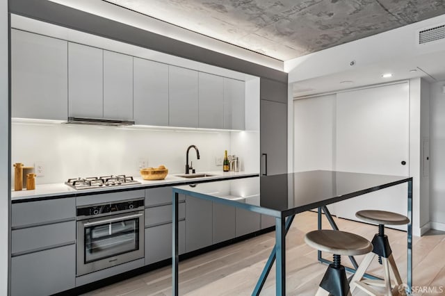 kitchen with gray cabinetry, a sink, appliances with stainless steel finishes, light wood-type flooring, and range hood