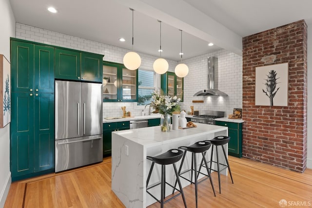 kitchen featuring pendant lighting, wall chimney range hood, appliances with stainless steel finishes, a center island, and light stone counters