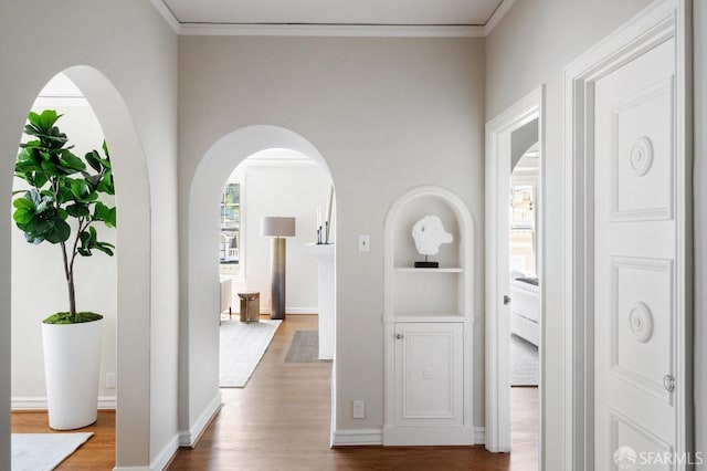 hallway with wood finished floors, built in features, arched walkways, baseboards, and ornamental molding