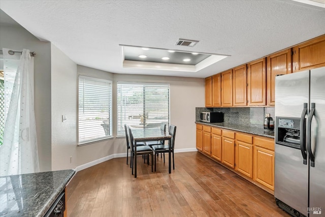 kitchen featuring a raised ceiling, wood-type flooring, appliances with stainless steel finishes, dark stone counters, and decorative backsplash