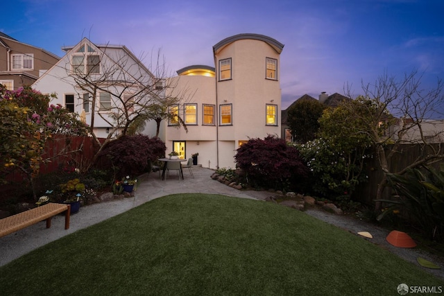 back of property at dusk featuring stucco siding, fence, a yard, and a patio area