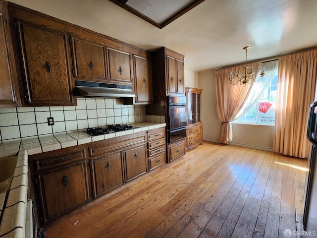 kitchen with tile countertops, black oven, decorative light fixtures, gas stovetop, and light hardwood / wood-style flooring