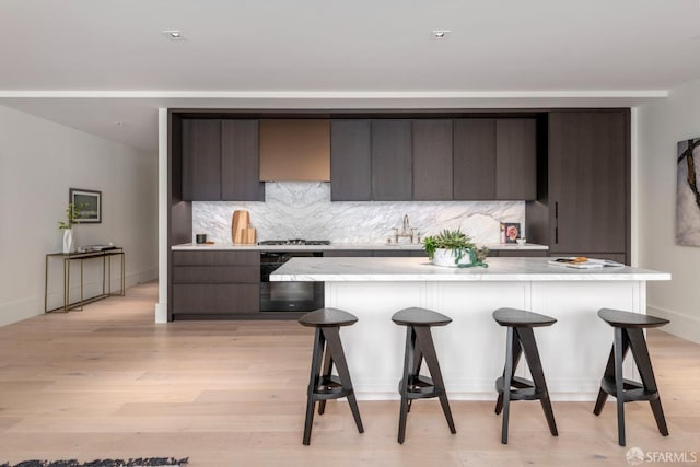 kitchen featuring light countertops, backsplash, light wood-style floors, dark brown cabinetry, and a kitchen bar