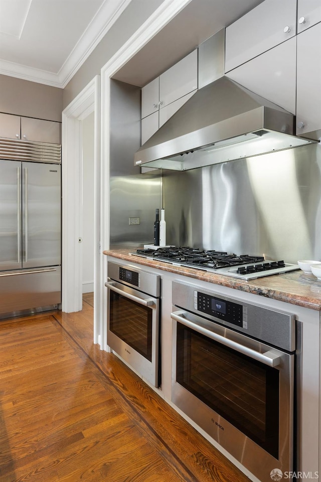 kitchen featuring appliances with stainless steel finishes, white cabinets, ornamental molding, hardwood / wood-style floors, and wall chimney range hood