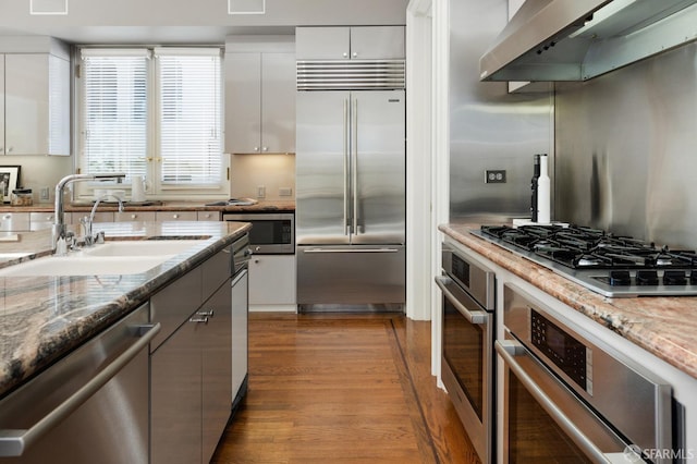 kitchen with exhaust hood, built in appliances, stone counters, and white cabinets