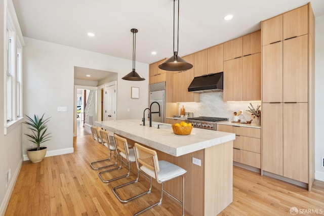 kitchen with modern cabinets, appliances with stainless steel finishes, light brown cabinetry, under cabinet range hood, and a kitchen bar