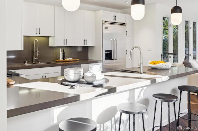 kitchen with a breakfast bar area, a sink, stainless steel built in fridge, white cabinets, and dark countertops