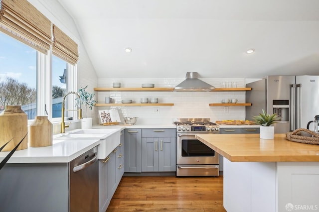 kitchen featuring open shelves, gray cabinets, vaulted ceiling, appliances with stainless steel finishes, and wall chimney exhaust hood