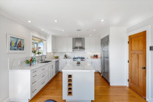 kitchen with light wood finished floors, wall chimney exhaust hood, light stone counters, stainless steel appliances, and a sink