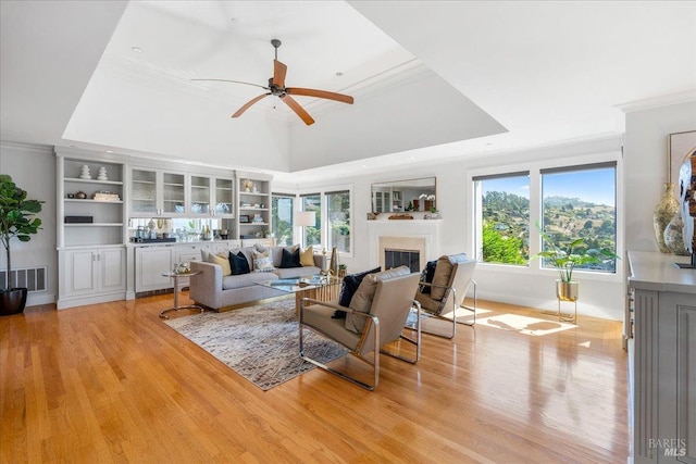 living area featuring visible vents, a glass covered fireplace, ornamental molding, a tray ceiling, and light wood-style floors