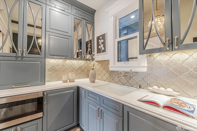 kitchen featuring a sink, light countertops, stainless steel microwave, backsplash, and glass insert cabinets
