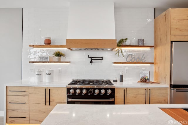 kitchen featuring custom exhaust hood, gas range oven, light brown cabinets, stainless steel refrigerator, and light stone countertops