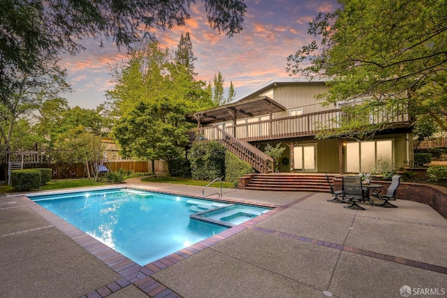 pool at dusk featuring stairs, a patio area, a wooden deck, a pool with connected hot tub, and fence