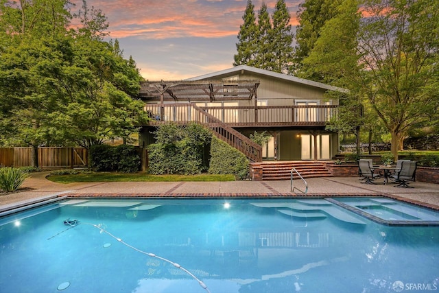 pool at dusk featuring fence, a deck, stairs, and a pool with connected hot tub