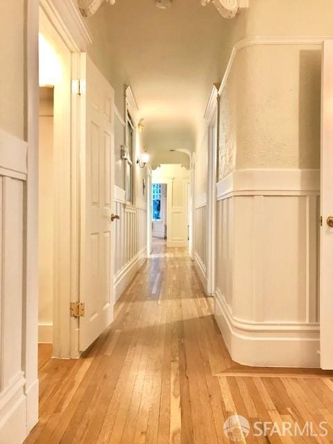 hall featuring light wood-style flooring, a decorative wall, and a wainscoted wall