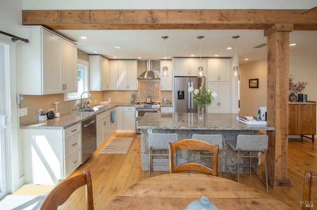 kitchen with light wood-style flooring, a sink, wall chimney exhaust hood, beamed ceiling, and appliances with stainless steel finishes