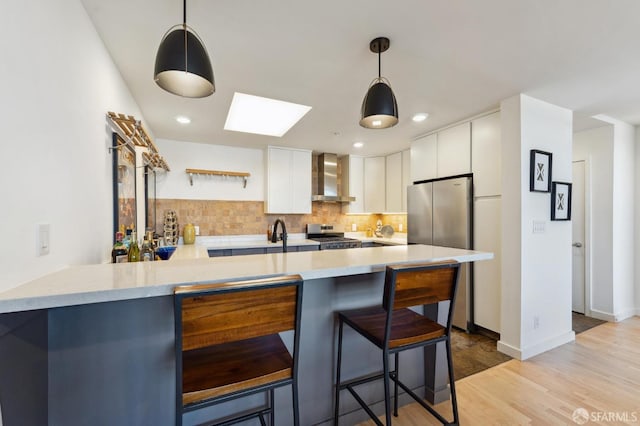 kitchen with kitchen peninsula, stainless steel appliances, wall chimney range hood, white cabinets, and hanging light fixtures