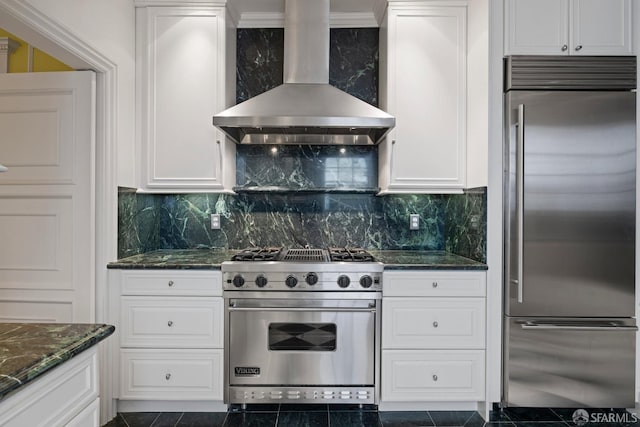 kitchen with high end appliances, white cabinetry, tasteful backsplash, dark stone counters, and wall chimney range hood