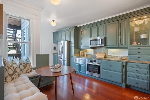 kitchen with tasteful backsplash, glass insert cabinets, appliances with stainless steel finishes, light stone counters, and dark wood-type flooring